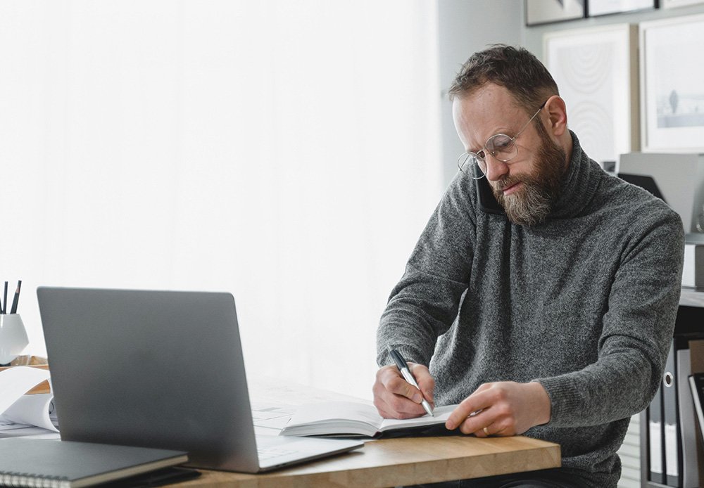 Focused man working at a desk with a laptop, writing in a notebook while talking on the phone, representing multitasking and managing important business tasks.