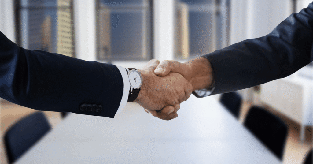 Two professionals in tailored suits shaking hands across a boardroom tab