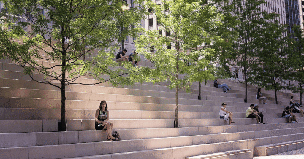 People sitting on tiered steps in an urban area with trees and high-rise buildings, illustrating local neighbourhood factors that influence US housing market fundamentals.